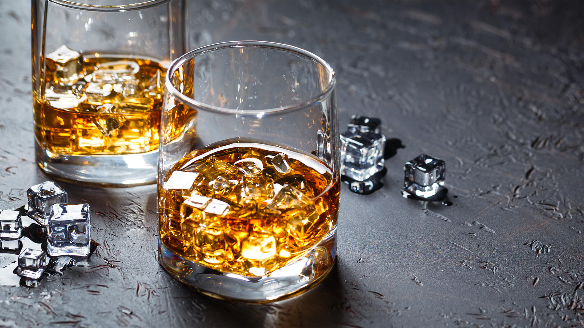 两个威士忌眼镜，在板岩表面上。每个玻璃杯中都有威士忌和冰，表面上有一些散落的冰块