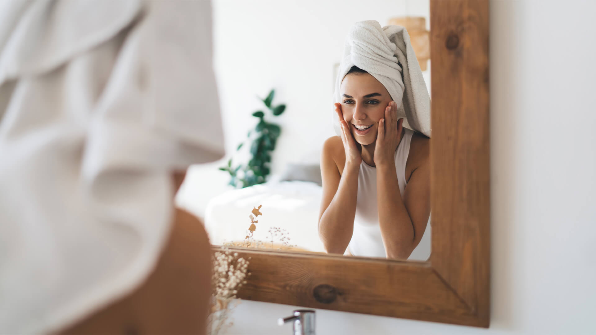 年轻的美丽女性，头上有毛巾，微笑着，镜面反射站在家里的浴室里。